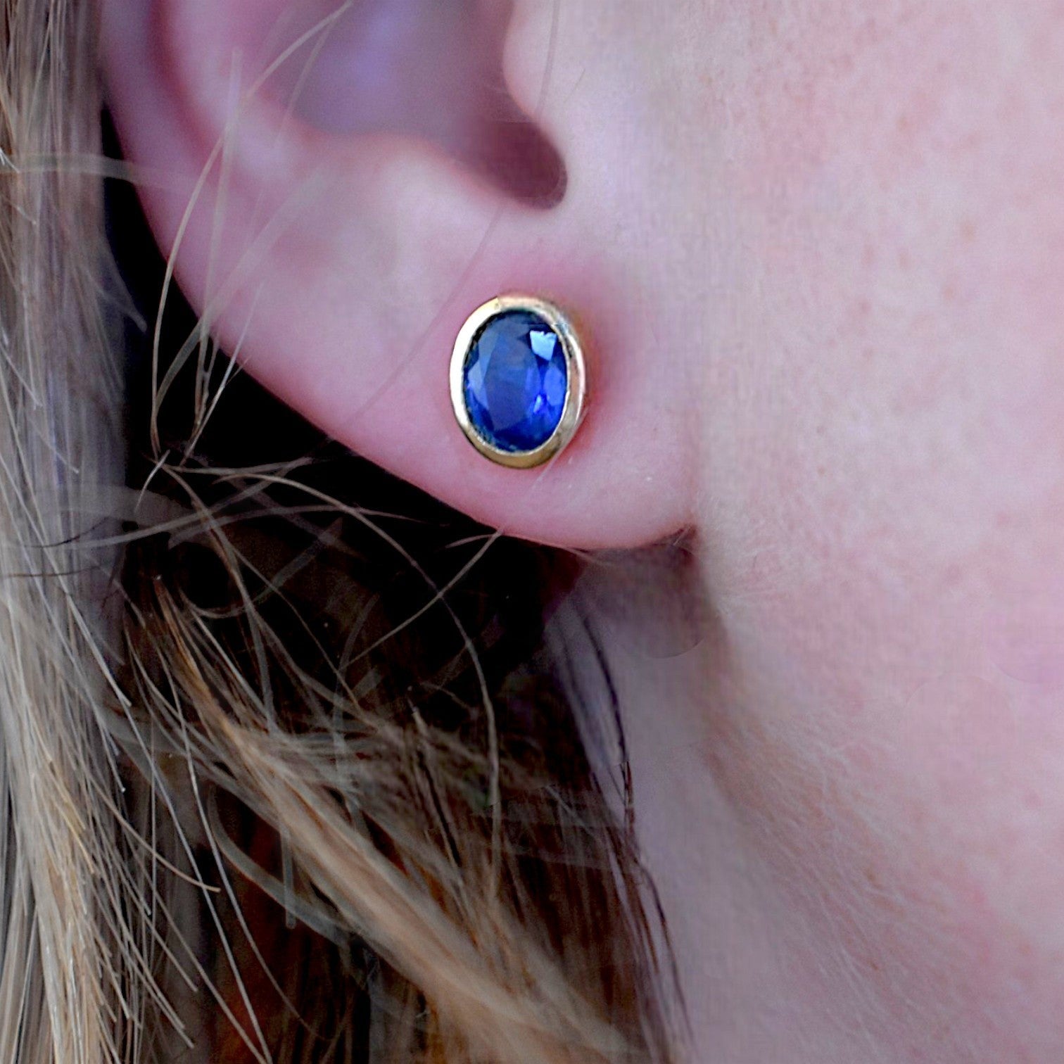 Natural blue sapphire oval stud earrings worn on an ear for scale and color vividness
